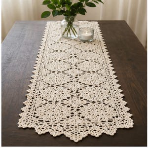 May include: Cream-colored, open-weave crochet table runner with a floral pattern and scalloped edges. The runner is displayed on a dark wood table, with a vase of greenery and a candle in the background.