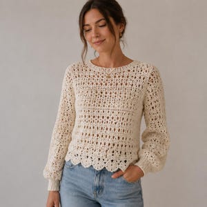 May include: Cream-colored crochet sweater with long sleeves and a scalloped hem. The sweater has an open, airy design with a round neckline and a relaxed fit. The sleeves are slightly puffed at the shoulders and fitted at the wrists. The model is wearing blue jeans.