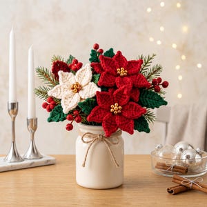 May include: A crochet floral arrangement featuring red and white poinsettia flowers with green leaves and red berries, accented with gold beads in the centers. The flowers are displayed in a white ceramic jar tied with a brown twine bow.