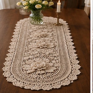 Op de afbeelding: Beige ovale tafelloper met een bloemen kanten ontwerp. De loper heeft driedimensionale rozen applicaties en een geschulpte rand. Een vaas met bloemen en een kandelaar staan op de tafel.