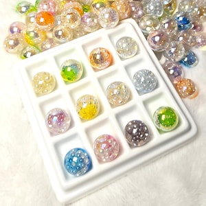 May include: A white plastic tray with twelve square compartments, each containing a single round, translucent bead filled with tiny white pearls. Additional loose, multi-colored, iridescent beads with pearl centers are scattered around the tray.