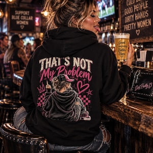 May include: A black hooded sweatshirt featuring a graphic on the back with the text That's Not My Problem in pink and white. The design includes a person wearing a cap, a heart, a crown, and a checkered pattern. The hoodie is made of black fabric.