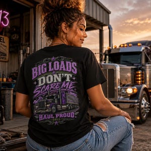 May include: A black t-shirt featuring a graphic on the back with purple and white text that reads BIG LOADS DON'T SCARE ME HAUL PROUD DP13. The design includes an illustration of a semi-truck with purple accents and decorative star elements.