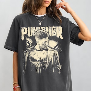 May include: Dark gray t-shirt featuring a graphic of "The Punisher". The design includes a portrait of a man and the Punisher skull logo. The text "The Punisher" is printed above the image in a distressed font.