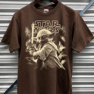 Może przedstawiać: Brązowa koszulka Star Wars z Yodą trzymającym miecz świetlny. Logo Star Wars jest wydrukowane nad Yodą. Koszulka jest z kr&oacute;tkim rękawem i wisi na drewnianym wieszaku.