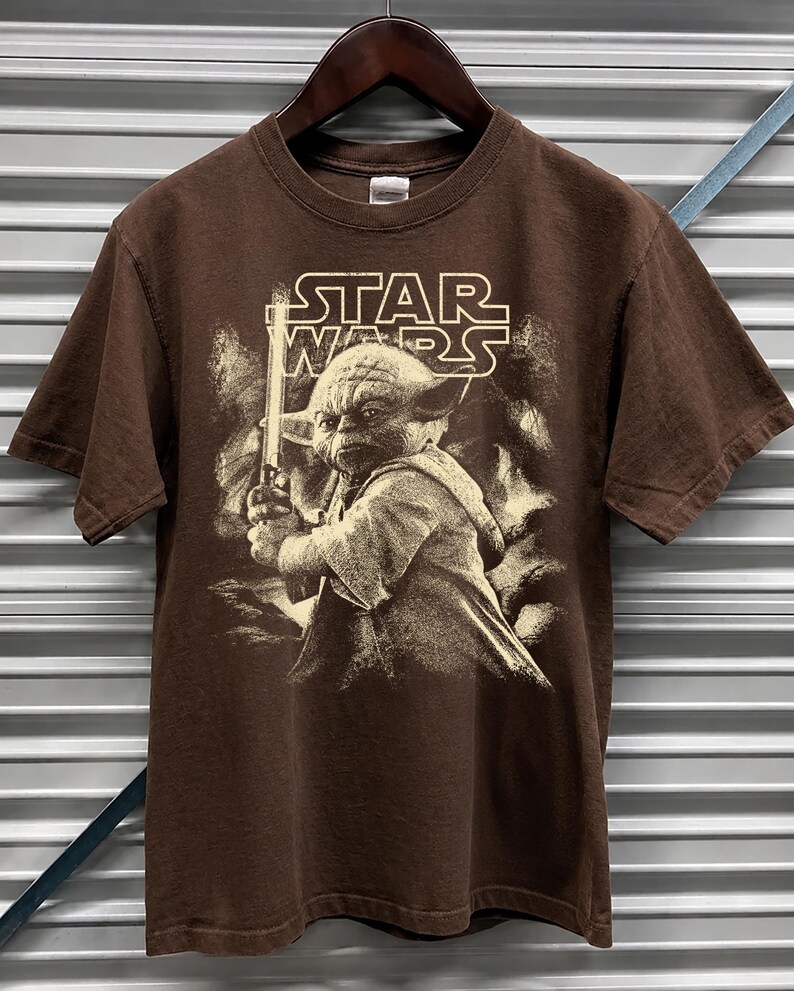 Może przedstawiać: Brązowa koszulka Star Wars z Yodą trzymającym miecz świetlny. Logo Star Wars jest wydrukowane nad Yodą. Koszulka jest z kr&oacute;tkim rękawem i wisi na drewnianym wieszaku.
