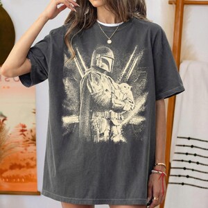 K&ouml;nnte beinhalten: Dunkelgraues T-Shirt mit einer Grafik eines Mandalorian-Charakters, der eine kleine Kreatur h&auml;lt. Das Design ist im Used-Vintage-Stil mit einer hellbeigen Farbe gehalten. Das Shirt hat kurze &Auml;rmel und einen Rundhalsausschnitt.