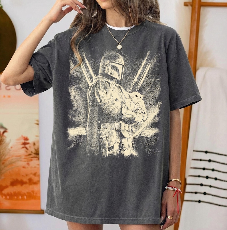 K&ouml;nnte beinhalten: Dunkelgraues T-Shirt mit einer Grafik eines Mandalorian-Charakters, der eine kleine Kreatur h&auml;lt. Das Design ist im Used-Vintage-Stil mit einer hellbeigen Farbe gehalten. Das Shirt hat kurze &Auml;rmel und einen Rundhalsausschnitt.