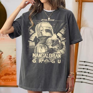 Puede incluir: Camiseta gris oscuro con un gr&aacute;fico de Star Wars: The Mandalorian. El dise&ntilde;o incluye el casco del Mandaloriano y Grogu. El texto "STAR WARS" est&aacute; encima de la imagen, y "THE MANDALORIAN AND GROGU" debajo.