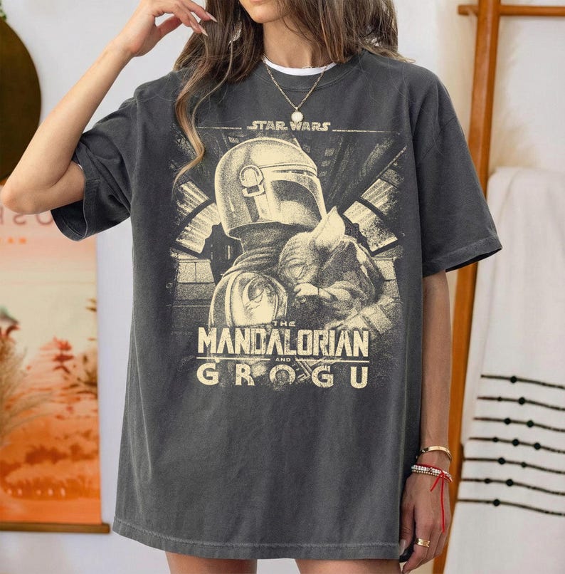 Puede incluir: Camiseta gris oscuro con un gr&aacute;fico de Star Wars: The Mandalorian. El dise&ntilde;o incluye el casco del Mandaloriano y Grogu. El texto "STAR WARS" est&aacute; encima de la imagen, y "THE MANDALORIAN AND GROGU" debajo.