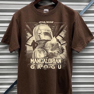 Puede incluir: Camiseta marr&oacute;n con un gr&aacute;fico en sepia de The Mandalorian y Grogu de Star Wars. El texto "The Mandalorian and Grogu" se muestra debajo de la imagen. La camiseta est&aacute; colgada en una percha de madera.