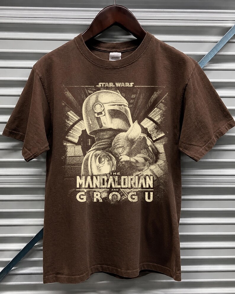 Puede incluir: Camiseta marr&oacute;n con un gr&aacute;fico en sepia de The Mandalorian y Grogu de Star Wars. El texto "The Mandalorian and Grogu" se muestra debajo de la imagen. La camiseta est&aacute; colgada en una percha de madera.