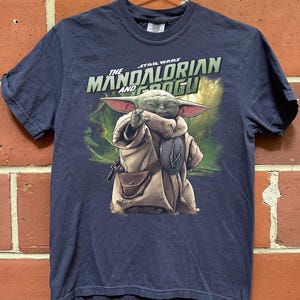 May include: Navy blue t-shirt with a graphic of Grogu from The Mandalorian. The image shows Grogu in a tan robe with a brown satchel, holding a silver emblem. The text "The Mandalorian and Grogu" is printed above the character.