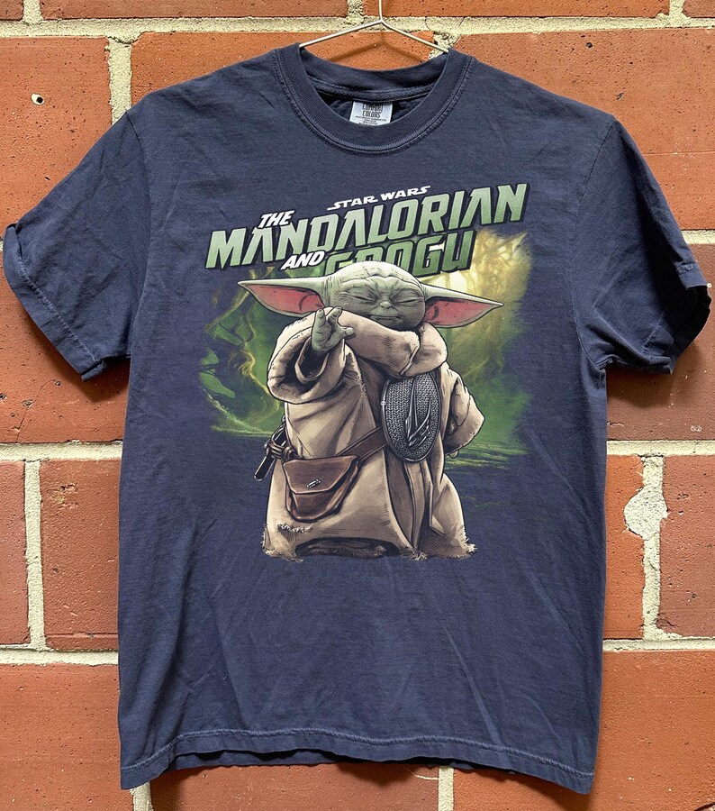 May include: Navy blue t-shirt with a graphic of Grogu from The Mandalorian. The image shows Grogu in a tan robe with a brown satchel, holding a silver emblem. The text "The Mandalorian and Grogu" is printed above the character.