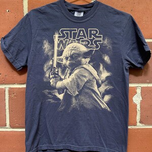 Może przedstawiać: Granatowa koszulka Star Wars z Yodą trzymającym miecz świetlny. Logo Star Wars jest wydrukowane nad Yodą. Grafika jest w stylu vintage. Koszulka wisi na metalowym wieszaku.
