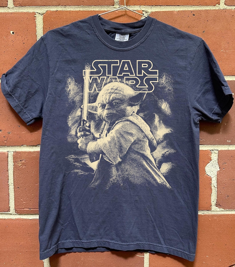 Może przedstawiać: Granatowa koszulka Star Wars z Yodą trzymającym miecz świetlny. Logo Star Wars jest wydrukowane nad Yodą. Grafika jest w stylu vintage. Koszulka wisi na metalowym wieszaku.