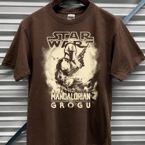 Puede incluir: Camiseta marr&oacute;n con un dise&ntilde;o gr&aacute;fico de la serie "The Mandalorian". El dise&ntilde;o incluye el logotipo de Star Wars, el personaje de Mandalorian y el texto "The Mandalorian and Grogu". La camiseta est&aacute; colgada de una percha de madera.