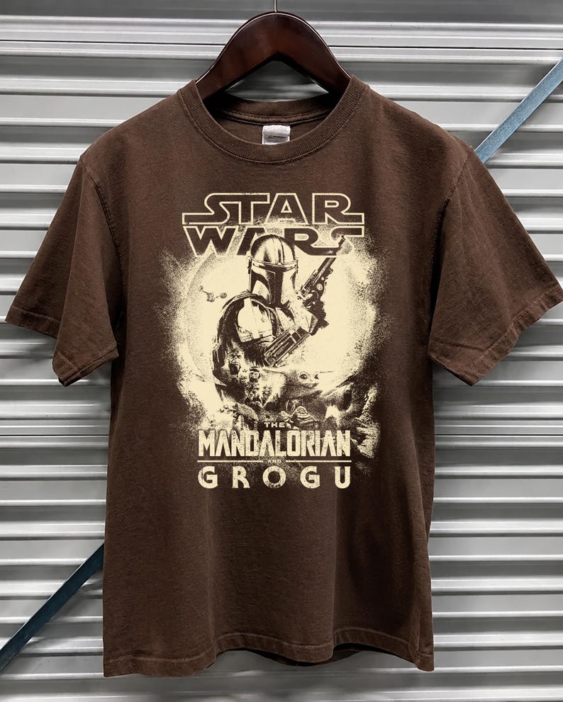 Puede incluir: Camiseta marr&oacute;n con un dise&ntilde;o gr&aacute;fico de la serie "The Mandalorian". El dise&ntilde;o incluye el logotipo de Star Wars, el personaje de Mandalorian y el texto "The Mandalorian and Grogu". La camiseta est&aacute; colgada de una percha de madera.