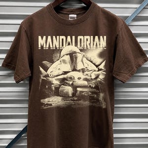 May include: A brown t-shirt with the word "MANDALORIAN" printed across the top. Below the text is a graphic of a helmeted figure and a small creature. The design has a faded, vintage appearance. The t-shirt hangs on a wooden hanger.