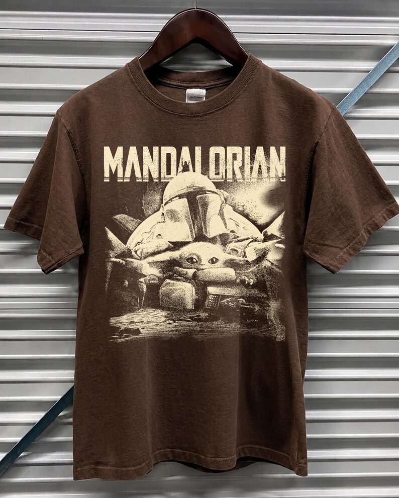 May include: A brown t-shirt with the word "MANDALORIAN" printed across the top. Below the text is a graphic of a helmeted figure and a small creature. The design has a faded, vintage appearance. The t-shirt hangs on a wooden hanger.