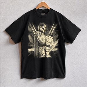K&ouml;nnte beinhalten: Schwarzes T-Shirt mit einer Grafik eines Mandalorian-Charakters, der eine kleine Kreatur h&auml;lt. Das Design ist in Beige- und Wei&szlig;t&ouml;nen gehalten und hat einen Used-Look. Das T-Shirt h&auml;ngt an einem Holzb&uuml;gel.