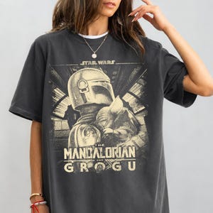 Puede incluir: Camiseta gris oscuro con un gr&aacute;fico de The Mandalorian y Grogu de Star Wars. El dise&ntilde;o incluye el texto "STAR WARS" encima de la imagen y "THE MANDALORIAN AND GROGU" debajo. La camiseta tiene mangas cortas y cuello redondo.