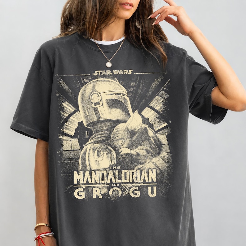 Puede incluir: Camiseta gris oscuro con un gr&aacute;fico de The Mandalorian y Grogu de Star Wars. El dise&ntilde;o incluye el texto "STAR WARS" encima de la imagen y "THE MANDALORIAN AND GROGU" debajo. La camiseta tiene mangas cortas y cuello redondo.