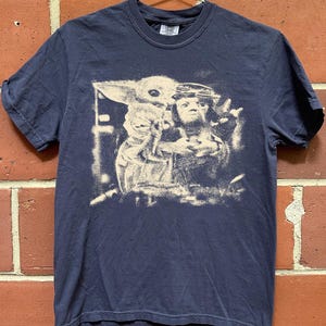 May include: Navy blue t-shirt with a distressed graphic of Grogu and Din Djarin from The Mandalorian. The image is in a vintage style. The shirt has short sleeves and a crew neck.