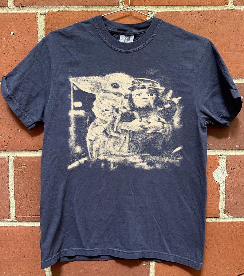 May include: Navy blue t-shirt with a distressed graphic of Grogu and Din Djarin from The Mandalorian. The image is in a vintage style. The shirt has short sleeves and a crew neck.