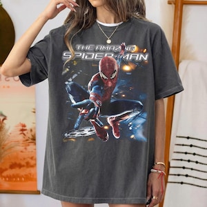 May include: Dark gray t-shirt featuring a graphic of Spider-Man from "The Amazing Spider-Man" movie. The superhero is in a dynamic pose, with the film's title printed above. The shirt has a vintage, worn look.