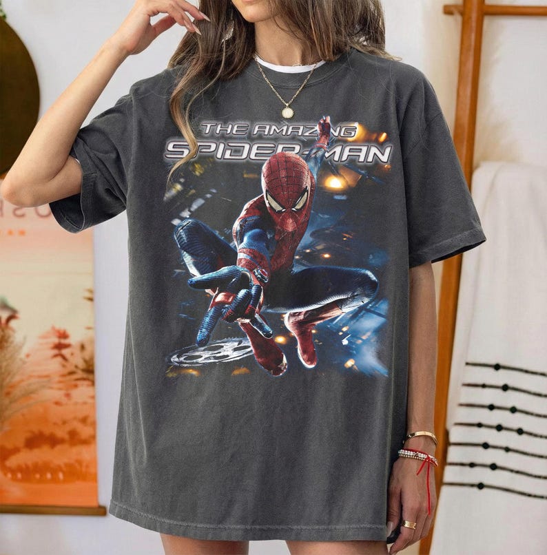 May include: Dark gray t-shirt featuring a graphic of Spider-Man from "The Amazing Spider-Man" movie. The superhero is in a dynamic pose, with the film's title printed above. The shirt has a vintage, worn look.