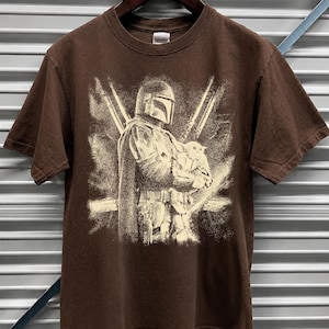 K&ouml;nnte beinhalten: Braunes T-Shirt mit einem Aufdruck, der eine Figur mit Helm zeigt, die eine kleine Kreatur h&auml;lt, mit zwei gekreuzten Lichtschwertern im Hintergrund. Das Design ist in Off-White gehalten. Das T-Shirt h&auml;ngt an einem Holzb&uuml;gel.