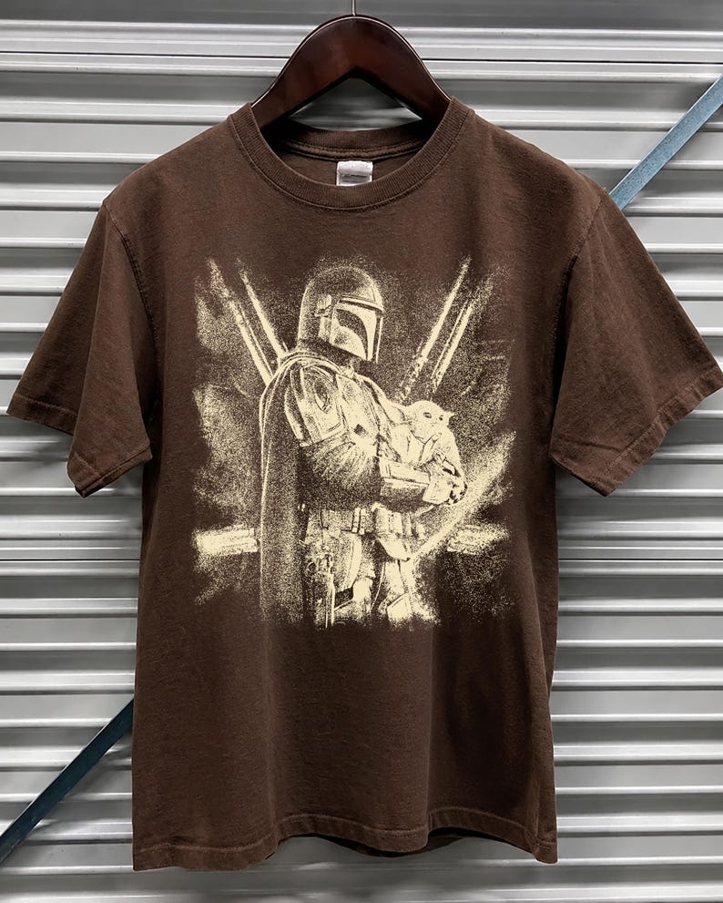 K&ouml;nnte beinhalten: Braunes T-Shirt mit einem Aufdruck, der eine Figur mit Helm zeigt, die eine kleine Kreatur h&auml;lt, mit zwei gekreuzten Lichtschwertern im Hintergrund. Das Design ist in Off-White gehalten. Das T-Shirt h&auml;ngt an einem Holzb&uuml;gel.