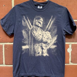 K&ouml;nnte beinhalten: Marineblaues T-Shirt mit einer Grafik von The Mandalorian, der Grogu h&auml;lt, mit zwei Lichtschwertern im Hintergrund. Das Design hat einen Used-Look im Vintage-Stil. Das T-Shirt h&auml;ngt an einem Metallb&uuml;gel vor einer Backsteinmauer.