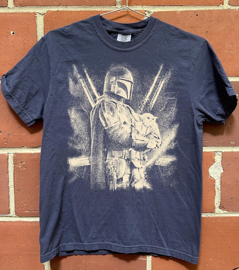 K&ouml;nnte beinhalten: Marineblaues T-Shirt mit einer Grafik von The Mandalorian, der Grogu h&auml;lt, mit zwei Lichtschwertern im Hintergrund. Das Design hat einen Used-Look im Vintage-Stil. Das T-Shirt h&auml;ngt an einem Metallb&uuml;gel vor einer Backsteinmauer.