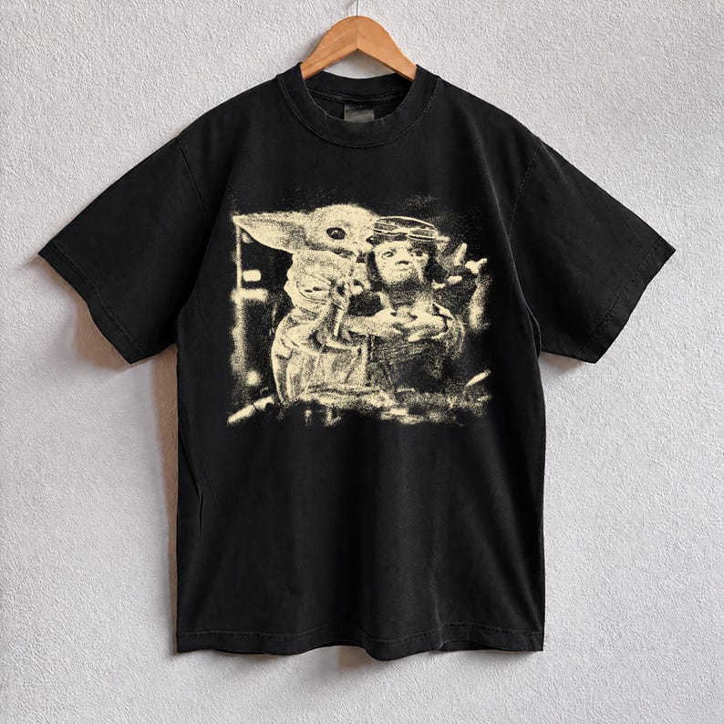 May include: Black t-shirt with a faded graphic of characters from a science fiction franchise. The design is in a light beige colour, giving it a vintage appearance. The t-shirt has short sleeves and a crew neck.