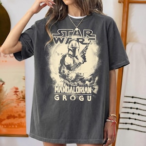 Puede incluir: Camiseta gris oscuro con un dise&ntilde;o de Star Wars. El dise&ntilde;o presenta el texto "STAR WARS" sobre una imagen de The Mandalorian, y el texto "THE MANDALORIAN GROGU" debajo. La camiseta tiene un corte holgado.