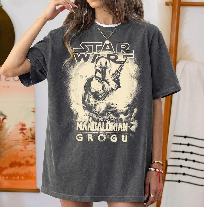 Puede incluir: Camiseta gris oscuro con un dise&ntilde;o de Star Wars. El dise&ntilde;o presenta el texto "STAR WARS" sobre una imagen de The Mandalorian, y el texto "THE MANDALORIAN GROGU" debajo. La camiseta tiene un corte holgado.