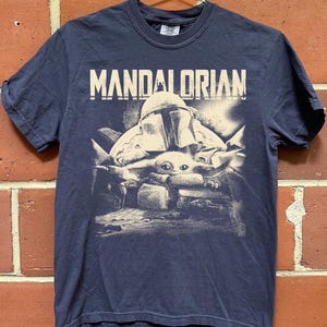 May include: Navy blue t-shirt with the word "MANDALORIAN" printed above a graphic of the Mandalorian and Grogu. The graphic is in white and grey tones. The shirt has short sleeves and a crew neck.