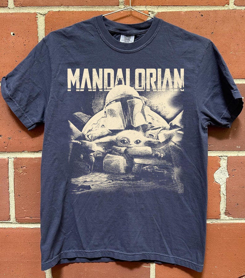 May include: Navy blue t-shirt with the word "MANDALORIAN" printed above a graphic of the Mandalorian and Grogu. The graphic is in white and grey tones. The shirt has short sleeves and a crew neck.