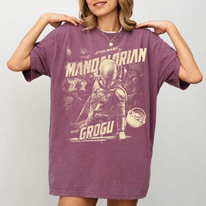 Pu&ograve; includere: Una maglietta color malva con una grafica della serie Star Wars, "The Mandalorian and Grogu". Il design include il personaggio di Mandalorian e Grogu, con il testo "The Mandalorian" e "Grogu" in stile vintage.