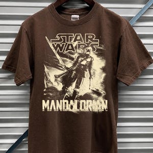 Pu&ograve; includere: T-shirt marrone con grafica di Star Wars. Il design include il testo "STAR WARS" e "MANDALORIAN" con l'immagine di un personaggio. La t-shirt &egrave; esposta su una gruccia di legno.