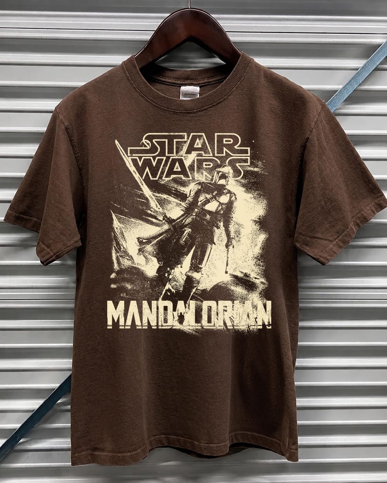 Pu&ograve; includere: T-shirt marrone con grafica di Star Wars. Il design include il testo "STAR WARS" e "MANDALORIAN" con l'immagine di un personaggio. La t-shirt &egrave; esposta su una gruccia di legno.