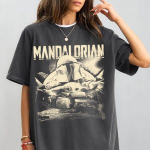 May include: Dark grey t-shirt with the word "MANDALORIAN" in off-white lettering. The graphic shows a helmeted figure and a small creature. The shirt has short sleeves and a crew neck.