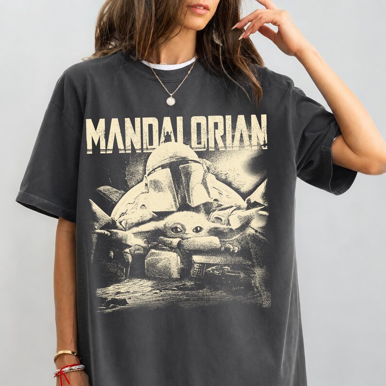 May include: Dark grey t-shirt with the word "MANDALORIAN" in off-white lettering. The graphic shows a helmeted figure and a small creature. The shirt has short sleeves and a crew neck.