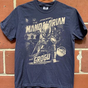 Pu&ograve; includere: T-shirt blu navy con una grafica di The Mandalorian e Grogu di Star Wars. Il design include il Mandaloriano con la sua armatura e Grogu nel suo trasportino. Il testo "The Mandalorian" e "Grogu" &egrave; stampato in stile vintage.