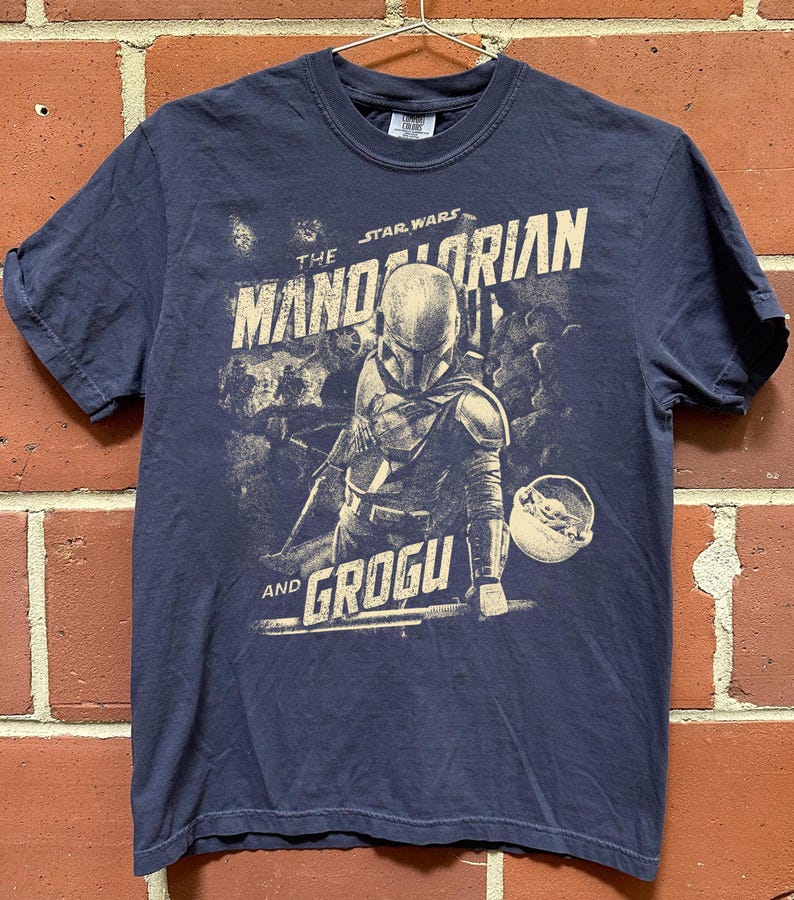 Pu&ograve; includere: T-shirt blu navy con una grafica di The Mandalorian e Grogu di Star Wars. Il design include il Mandaloriano con la sua armatura e Grogu nel suo trasportino. Il testo "The Mandalorian" e "Grogu" &egrave; stampato in stile vintage.