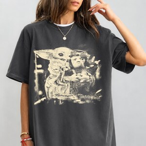 May include: Dark grey t-shirt with a sepia-toned graphic of a character holding a small, green-skinned creature. The design has a distressed, vintage appearance. The shirt has short sleeves and a crew neck.