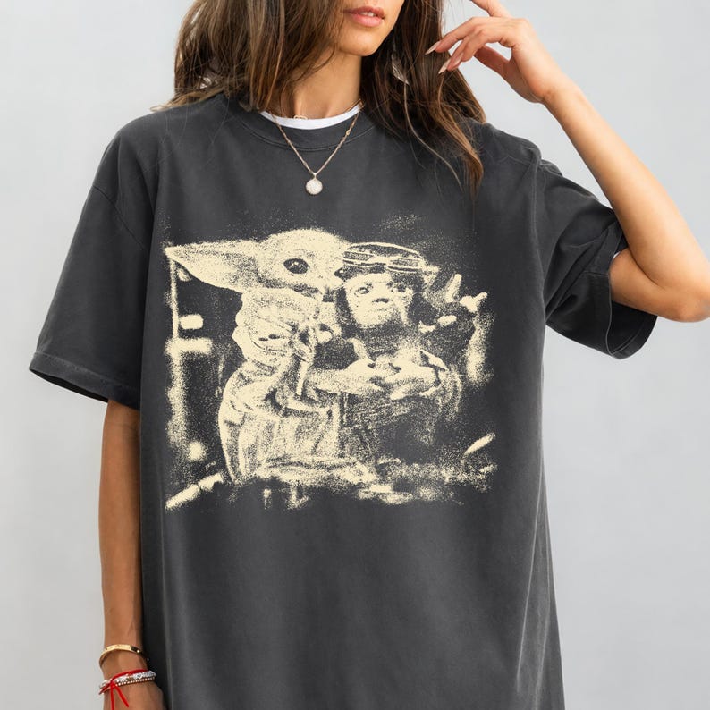 May include: Dark grey t-shirt with a sepia-toned graphic of a character holding a small, green-skinned creature. The design has a distressed, vintage appearance. The shirt has short sleeves and a crew neck.