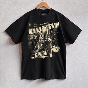 Pu&ograve; includere: T-shirt nera con grafica vintage di The Mandalorian e Grogu di Star Wars. Il design include il testo "THE MANDALORIAN" e "AND GROGU" in stile vintage, con un'immagine di The Mandalorian e Grogu.